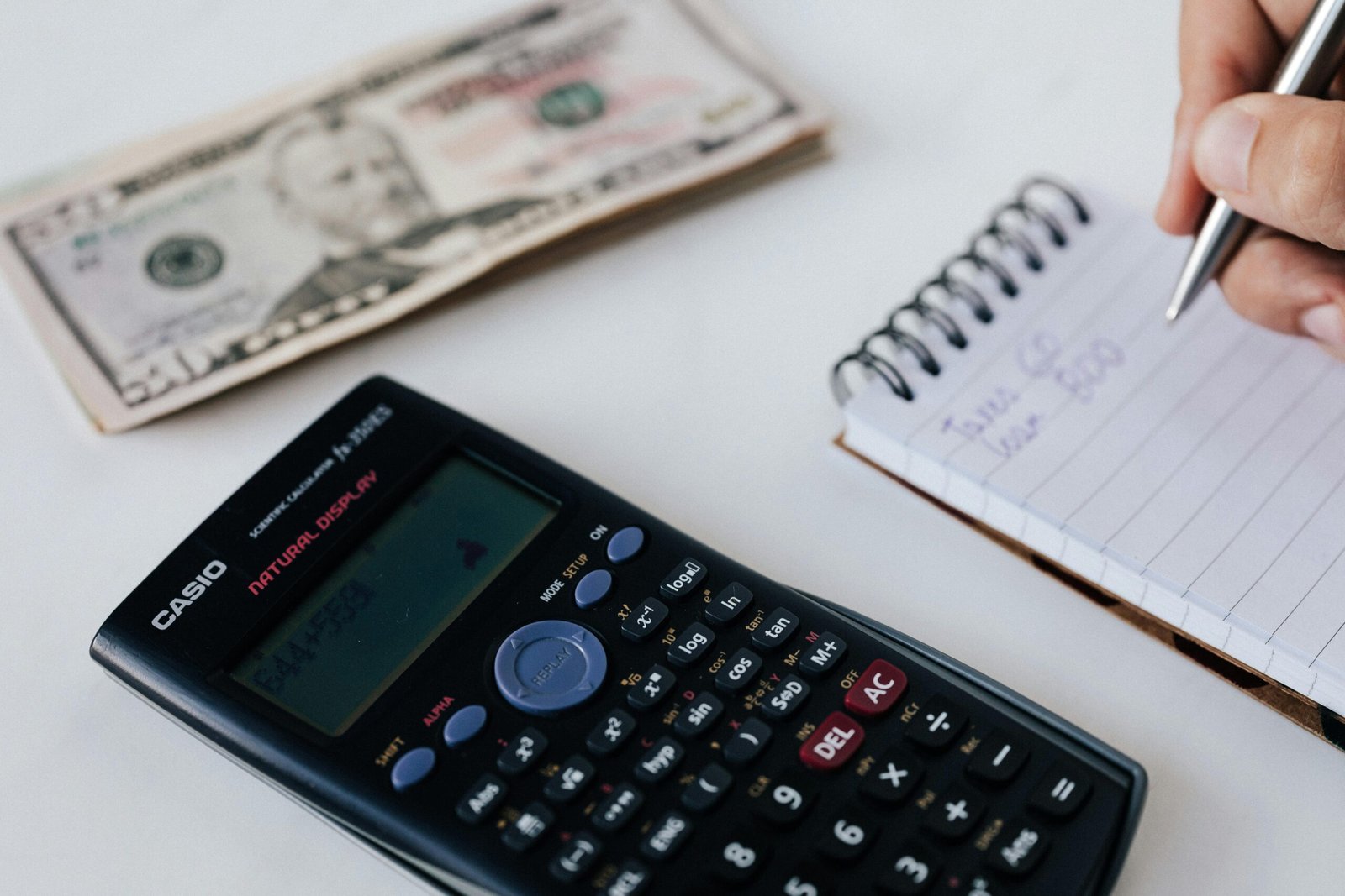 Home Close-up of a calculator, US dollar bill, and a hand writing in a notebook, symbolizing financial planning.